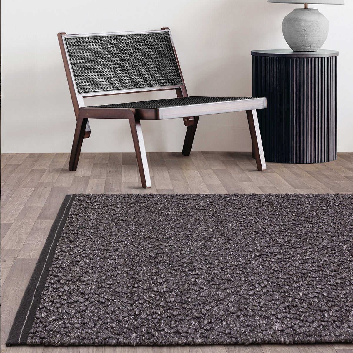 Dark gray textured rug on a wooden floor with a woven chair and side table in the background.