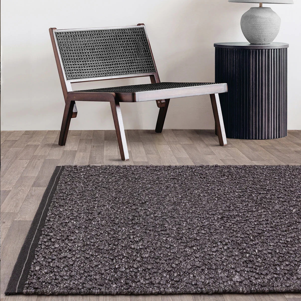 Dark gray textured rug on a wooden floor with a woven chair and side table in the background.