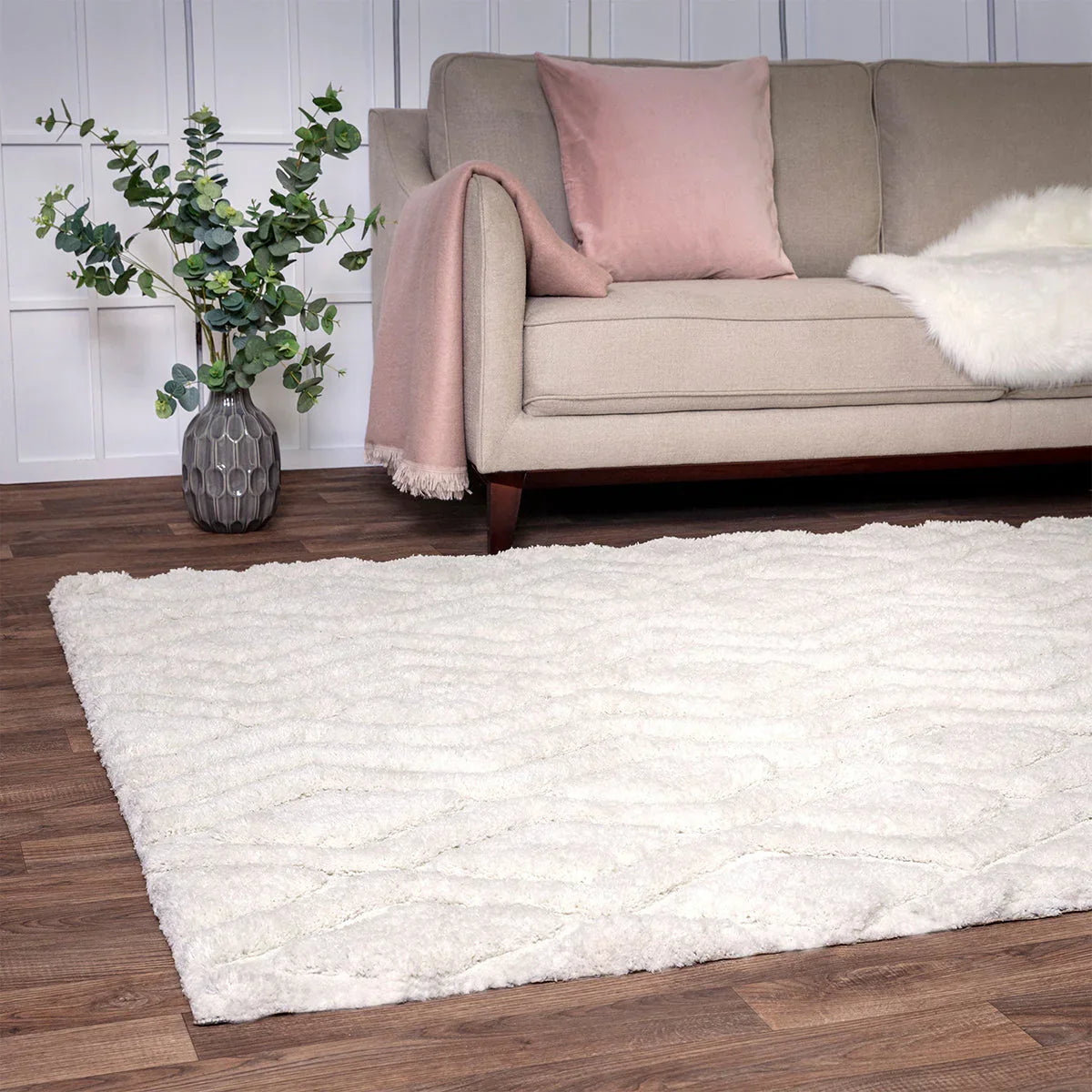 Beige sofa with pink pillow and blanket on a white rug in a room with wooden flooring.