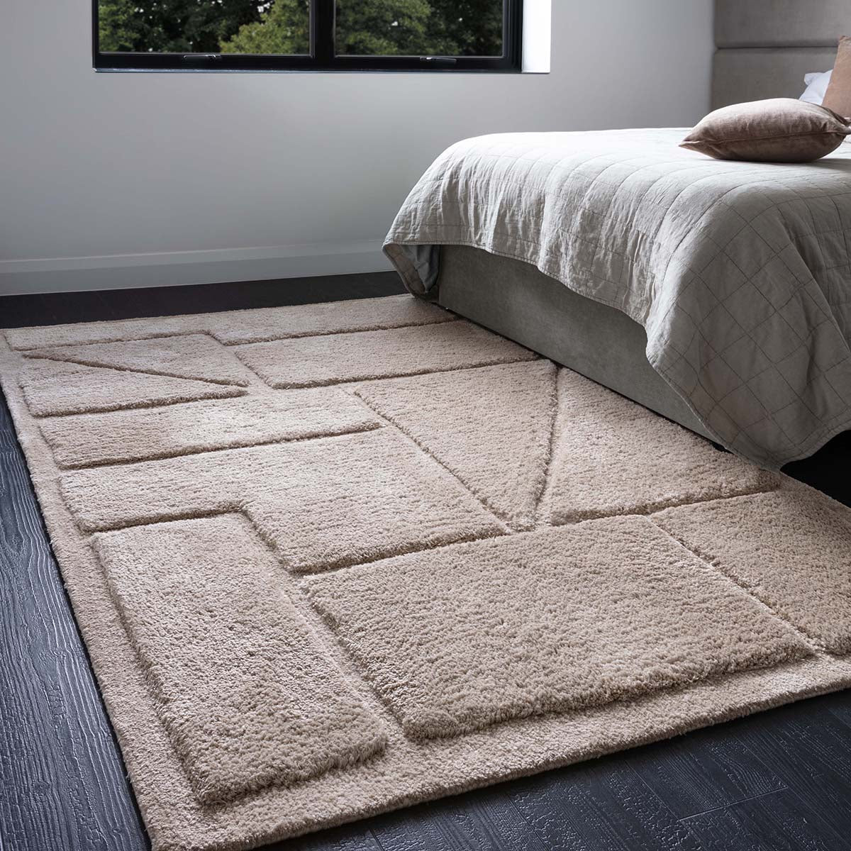 Beige geometric patterned rug in a bedroom setting with a bed and window.