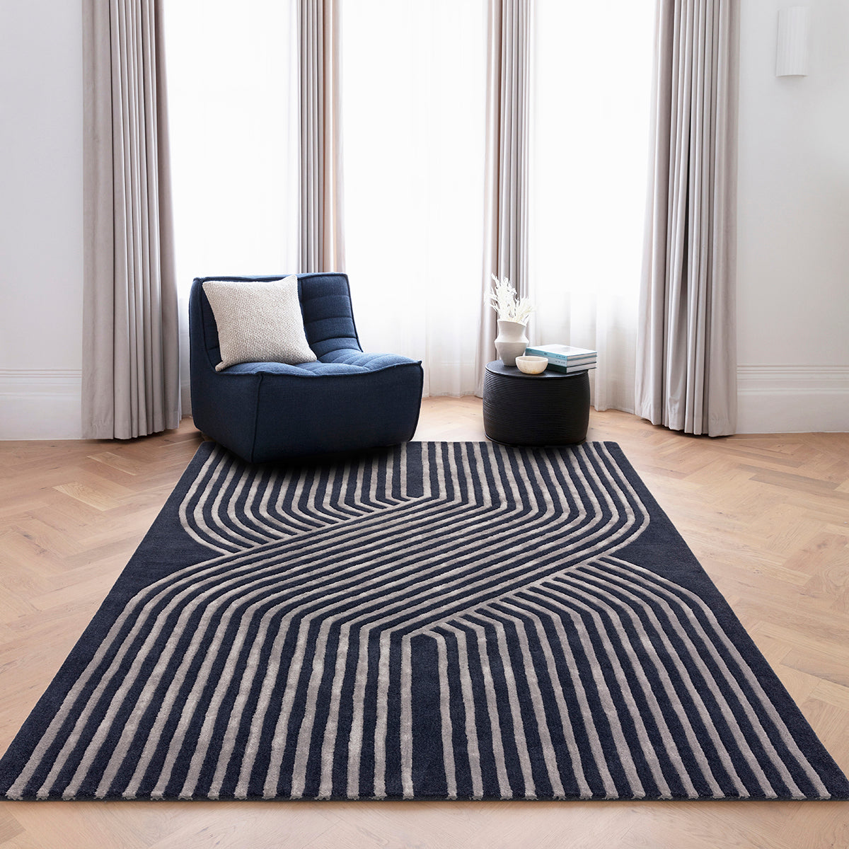 Navy blue armchair with a white pillow on a navy blue rug with white stripes in a room with large windows and curtains.