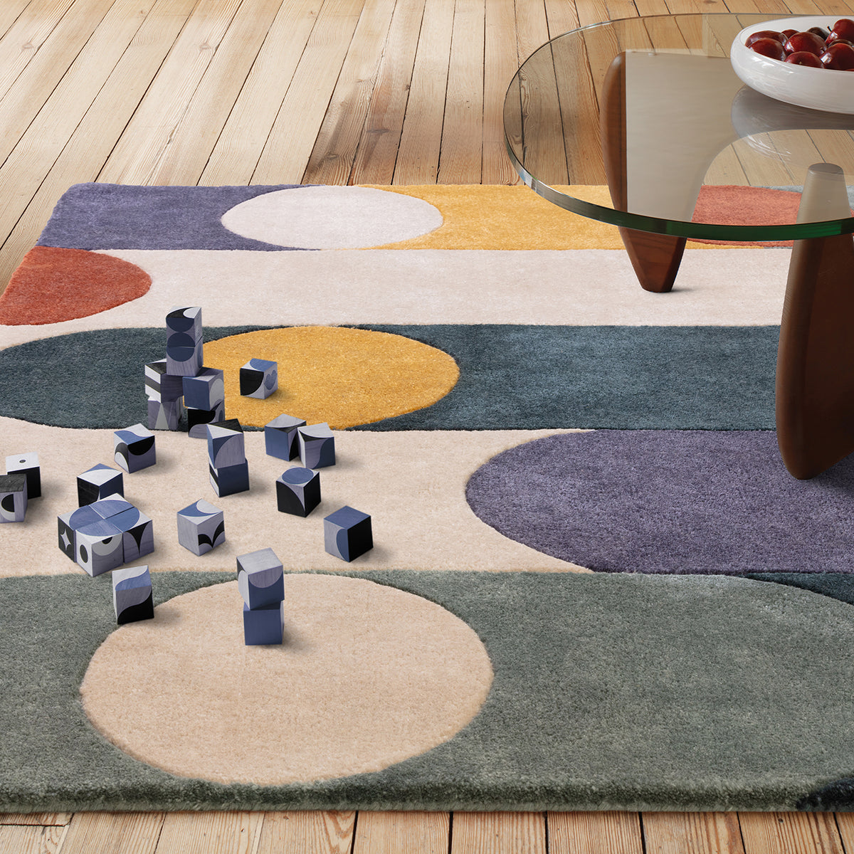 Colorful abstract rug with geometric shapes on a wooden floor, featuring a glass coffee table and decorative bowl.