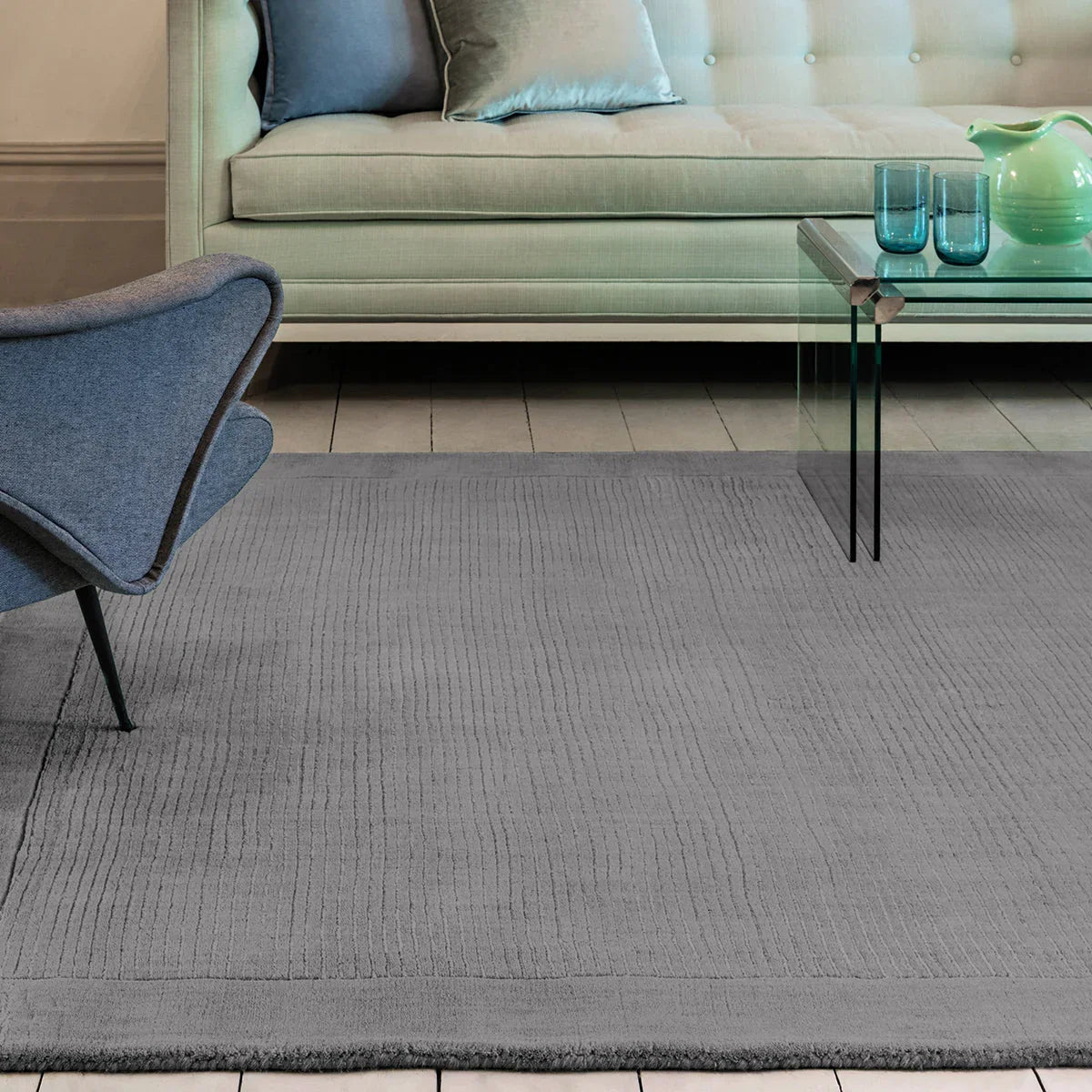 Gray rug in a living room with a blue chair, glass coffee table, and beige sofa.
