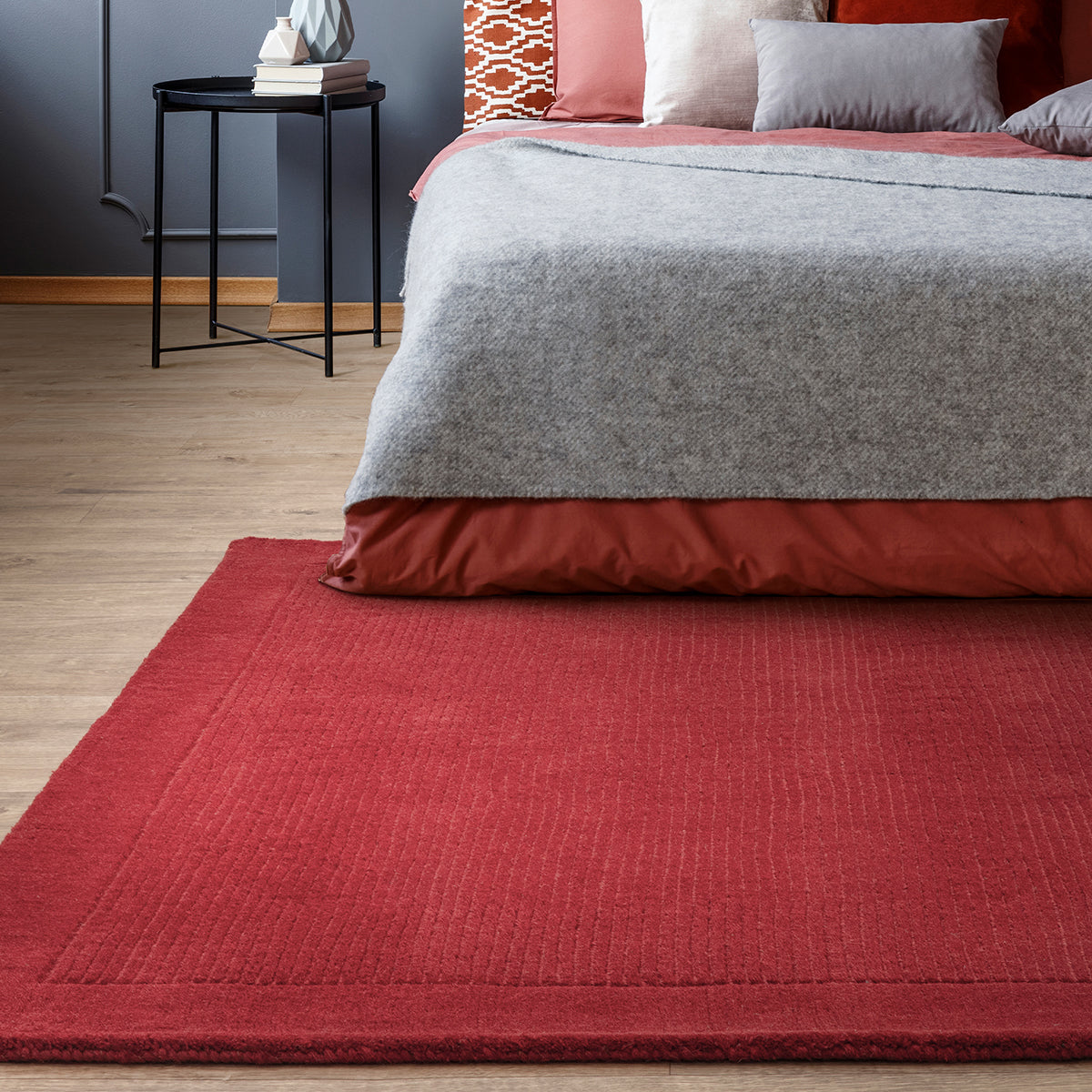 Red rug in front of a bed with gray and red bedding in a bedroom setting.