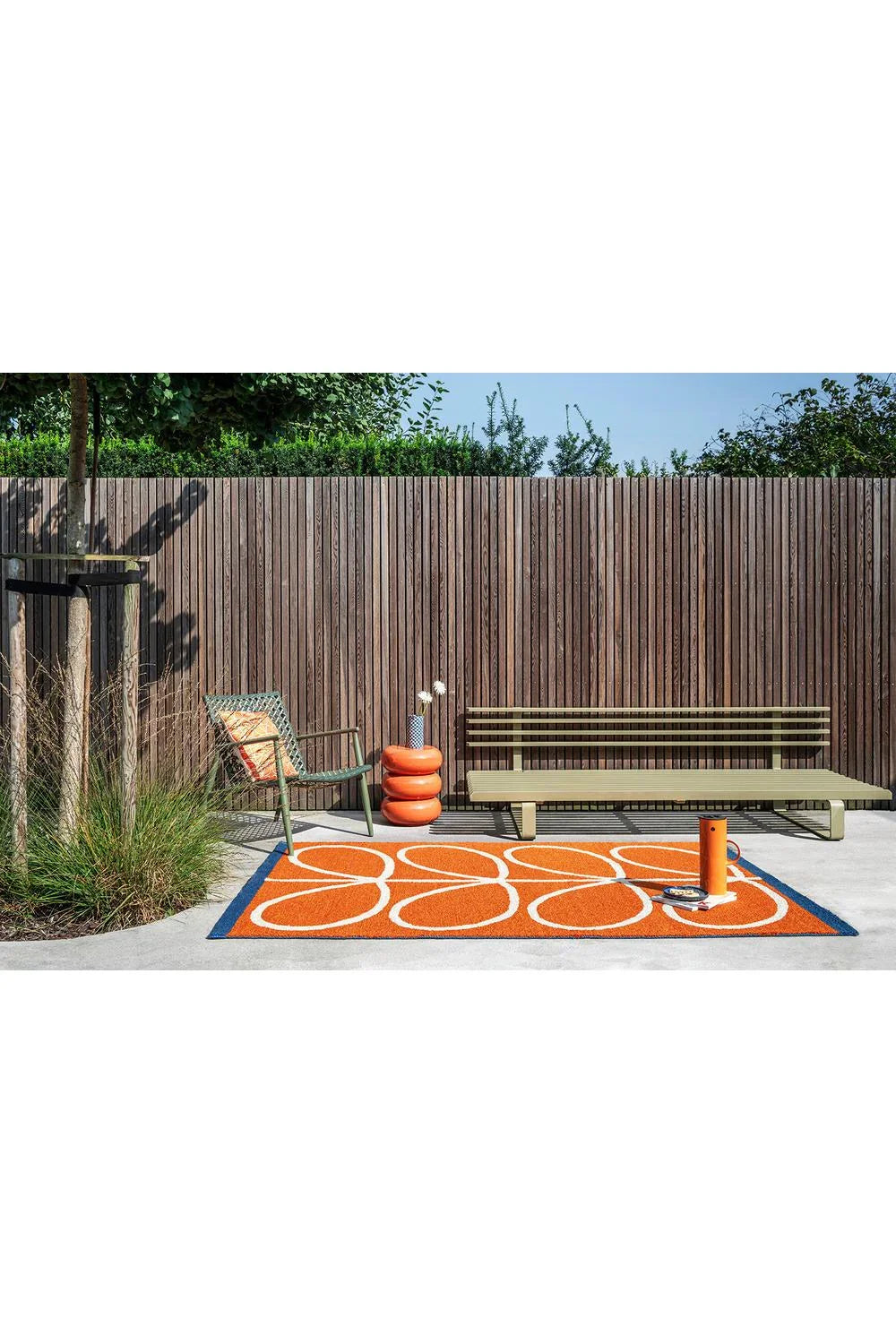 Orla Kiely Linear Stem Orange Outdoor Rug featuring circular orange and white motif with blue border on grey patio, power-loomed polypropylene