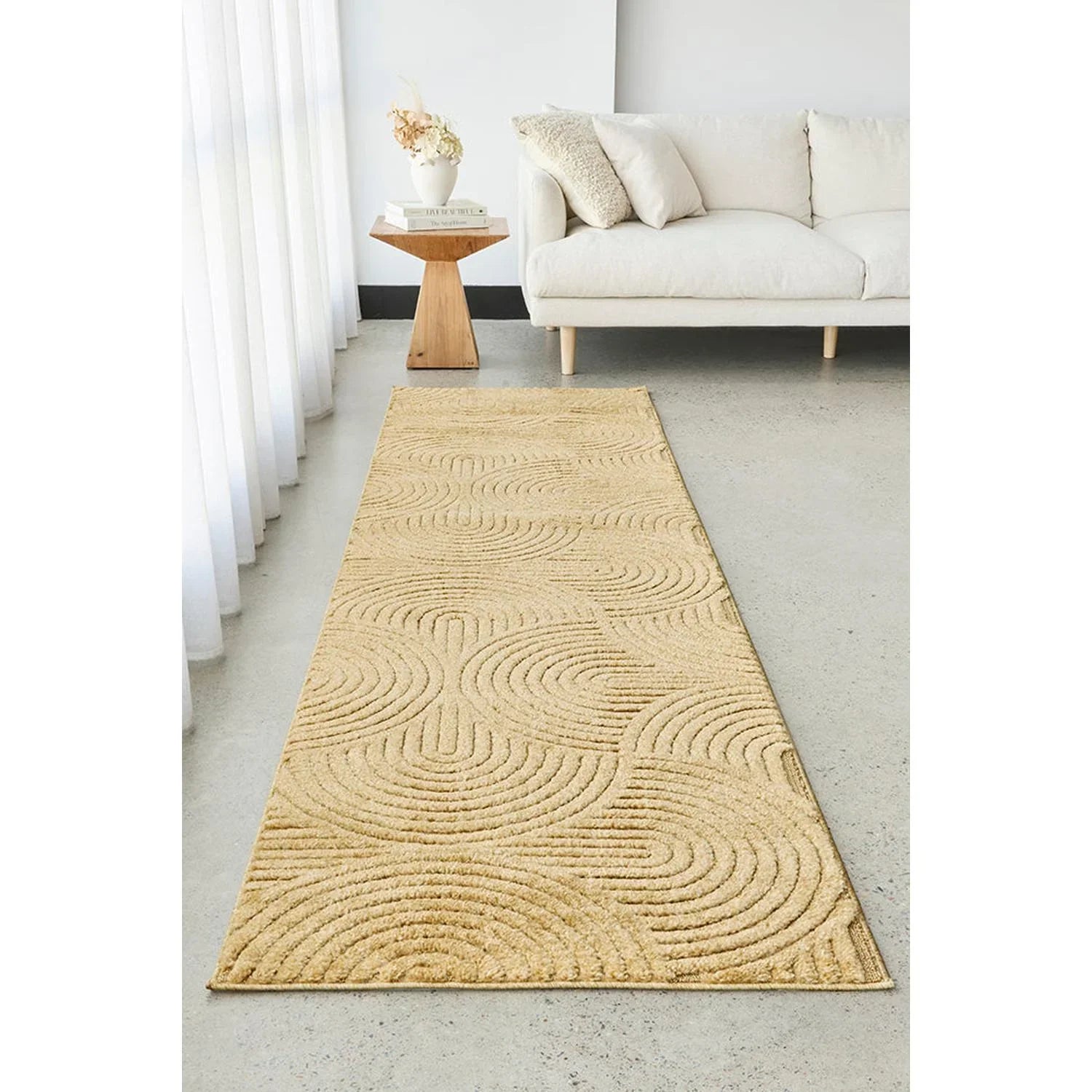 Beige patterned rug on a light gray floor in a living room setting with a white sofa and wooden side table.