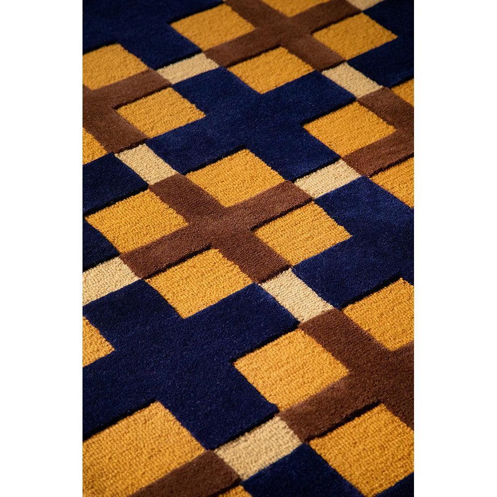 Brink & Campman Decor Milano rug featuring geometric grid pattern in navy blue, golden ochre, brown and beige on dark backdrop, hand-tufted wool